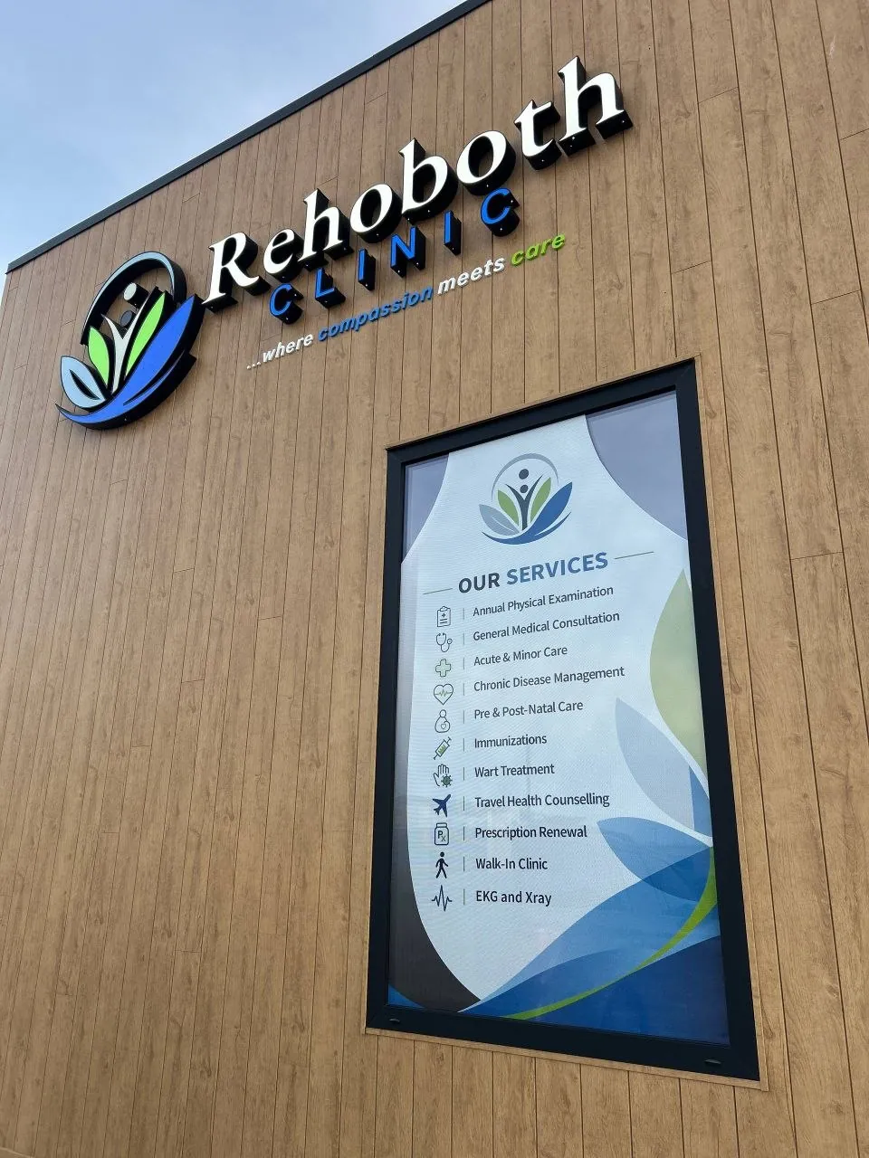 Rehoboth Clinic lit exterior sign with dimensional letters and logo above a window graphic listing clinic services